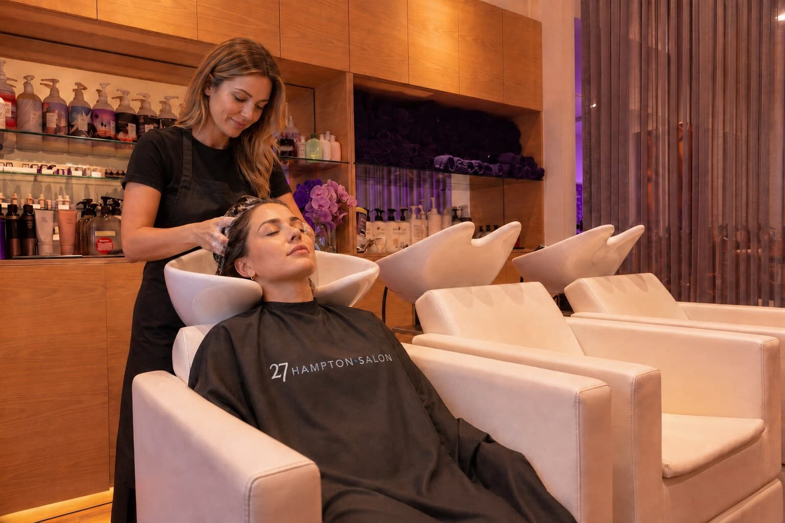 A stylist at 27 Hampton Salon shampooing a client reclined at one of the wash stations, with warm wood shelving in the background